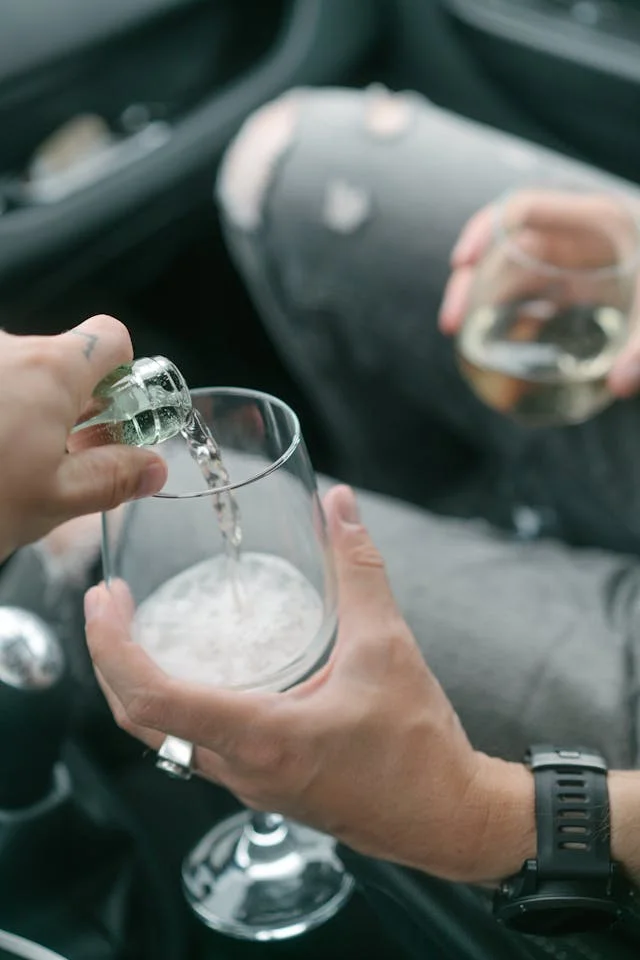 Two friends enjoying drinks inside a car, pouring and holding wine glasses.
